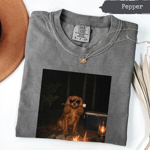 May include: A pepper-coloured crew neck t-shirt featuring a graphic of a brown dog wearing sunglasses and holding a marshmallow on a stick. The dog is positioned in a dark forest setting near a campfire, a cooler, and a lit lantern. The shirt has a Comfort Colors brand label at the inner neckline.