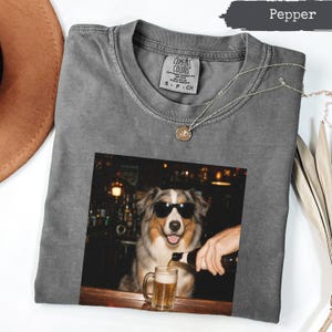 May include: A charcoal grey crew neck t-shirt featuring a graphic print of an Australian Shepherd dog wearing black sunglasses, sitting at a bar, and having a beer poured into a glass mug. The shirt has a Comfort Colors brand label at the neck.