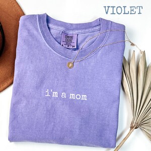 May include: A violet Comfort Colors t-shirt with the embroidered text "i'm a mom". A gold necklace with a pendant is draped over the shirt. A brown hat and dried plant are in the background.