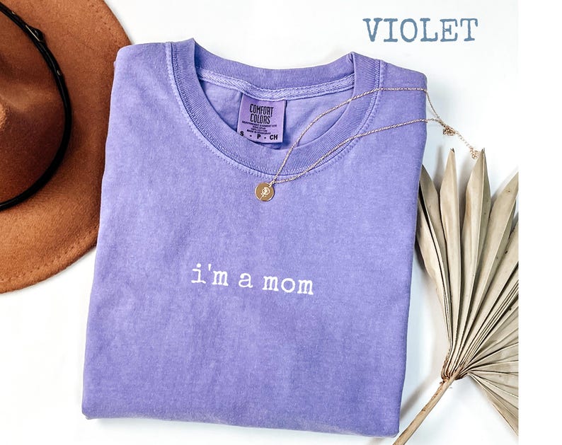 May include: A violet Comfort Colors t-shirt with the embroidered text "i'm a mom". A gold necklace with a pendant is draped over the shirt. A brown hat and dried plant are in the background.
