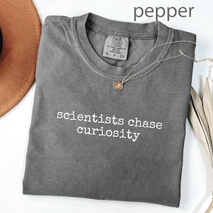 May include: A gray t-shirt with the text "scientists chase curiosity" in white. A gold necklace with a pendant rests on the shirt. The shirt is folded and next to a brown hat and dried plant.