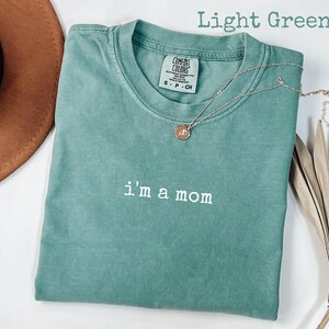 May include: Light green t-shirt with the embroidered text "i'm a mom". A gold-coloured necklace with a pendant rests on the shirt. The shirt is folded, and a brown hat is partially visible in the background.