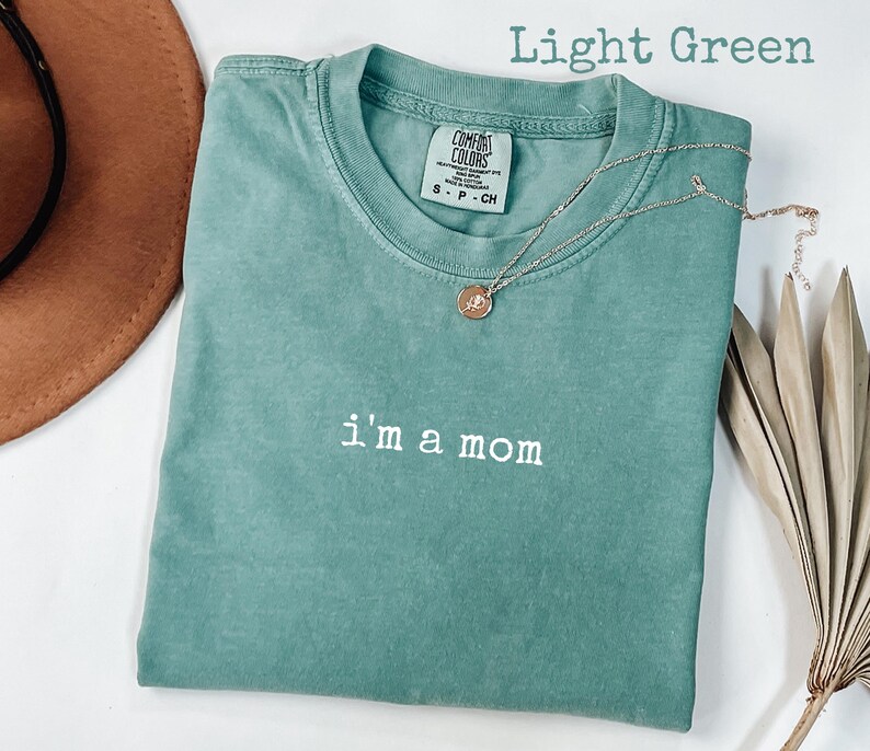 May include: Light green t-shirt with the embroidered text "i'm a mom". A gold-coloured necklace with a pendant rests on the shirt. The shirt is folded, and a brown hat is partially visible in the background.