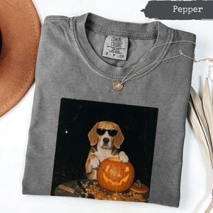 Peut inclure: Un t-shirt à col rond couleur poivre avec une impression graphique représentant un chien beagle portant des lunettes de soleil noires et sculptant une citrouille d'Halloween. Le t-shirt porte une étiquette Comfort Colors au col.