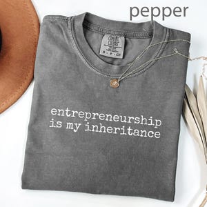 May include: A gray t-shirt with the text "entrepreneurship is my inheritance" in white. A gold necklace with a pendant is draped across the shirt. The shirt is folded, and a brown hat is partially visible in the background.