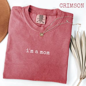 May include: Crimson t-shirt with the text "I'm a mom" in white. A gold-coloured necklace with a pendant is draped on the shirt. The shirt is a Comfort Colors brand, size S-P-CH.