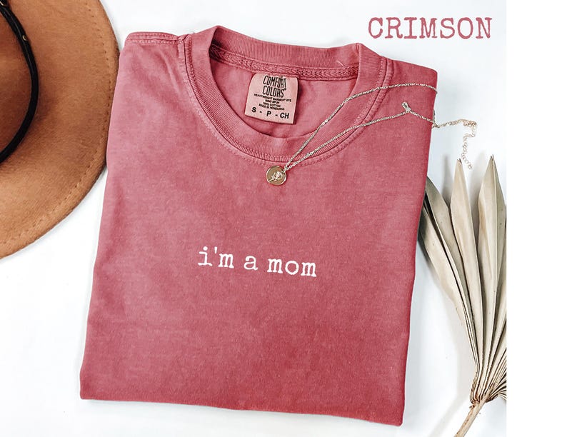 May include: Crimson t-shirt with the text "I'm a mom" in white. A gold-coloured necklace with a pendant is draped on the shirt. The shirt is a Comfort Colors brand, size S-P-CH.