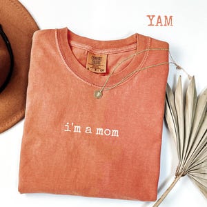 May include: A coral-coloured Comfort Colors t-shirt with the embroidered phrase "i'm a mom." A gold necklace with a pendant rests on the shirt. A brown hat and dried palm frond are in the background. The word "YAM" is printed in the upper right corner.