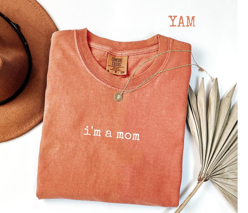 May include: A coral-coloured Comfort Colors t-shirt with the embroidered phrase "i'm a mom." A gold necklace with a pendant rests on the shirt. A brown hat and dried palm frond are in the background. The word "YAM" is printed in the upper right corner.