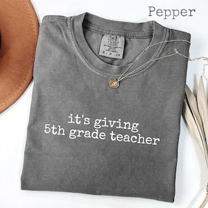 Op de afbeelding: Grijs T-shirt met de witte tekst "it's giving 5th grade teacher". Een zilveren ketting met een hanger ligt op het shirt. Een bruine hoed en een gedroogde plant staan op de achtergrond.