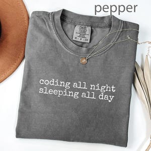 Op de afbeelding: Een grijs t-shirt met de tekst "coding all night sleeping all day" in het wit. Het shirt is opgevouwen en heeft een ronde halslijn. Een gouden ketting met een hanger ligt op het shirt. Een bruine hoed en een gedroogde plant staan op de achtergrond.