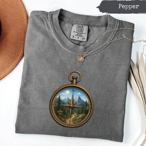 May include: A pepper-coloured short-sleeve t-shirt featuring a graphic of a round, bronze-toned compass. The compass face displays a detailed landscape with snow-capped mountains, pine trees, a winding path, and a sun in a blue sky. The shirt has a crew neckline and a Comfort Colors brand label at the inner collar.