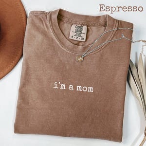 May include: Espresso-coloured t-shirt with the phrase "i'm a mom" in white lettering. The shirt has a crew neck and short sleeves. A silver necklace with a small pendant is draped across the shirt. The shirt is made of a soft material.