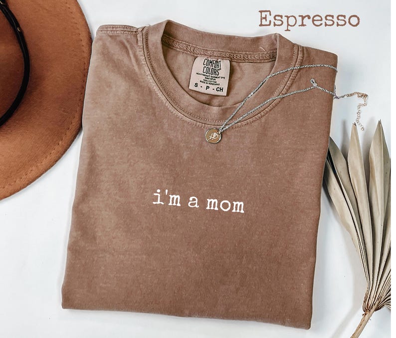 May include: Espresso-coloured t-shirt with the phrase "i'm a mom" in white lettering. The shirt has a crew neck and short sleeves. A silver necklace with a small pendant is draped across the shirt. The shirt is made of a soft material.