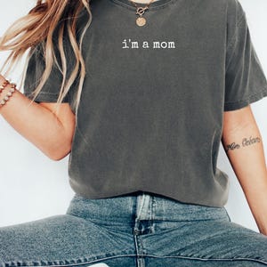 May include: Dark grey t-shirt with the text "i'm a mom" in white. The shirt has a casual style with short sleeves. The model is wearing blue jeans and a gold necklace.