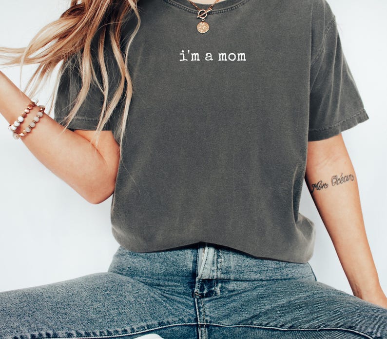 May include: Dark grey t-shirt with the text "i'm a mom" in white. The shirt has a casual style with short sleeves. The model is wearing blue jeans and a gold necklace.