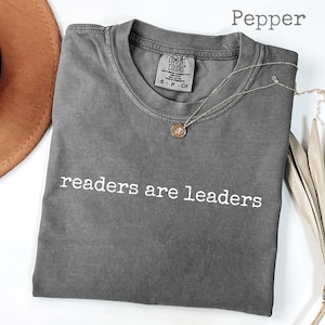 Puede incluir: Una camiseta gris con el texto blanco "readers are leaders". La camiseta está doblada y tiene cuello redondo. Un collar dorado con un colgante está sobre la camiseta. Un sombrero marrón y una planta seca están en el fondo.