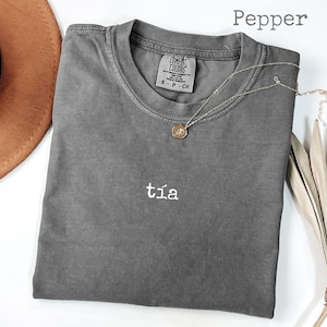 May include: A gray Comfort Colors t-shirt with the word "tía" embroidered in white. A gold pendant necklace rests on the shirt. The word "Pepper" is printed in the upper right corner. A brown hat and dried plant are in the background.