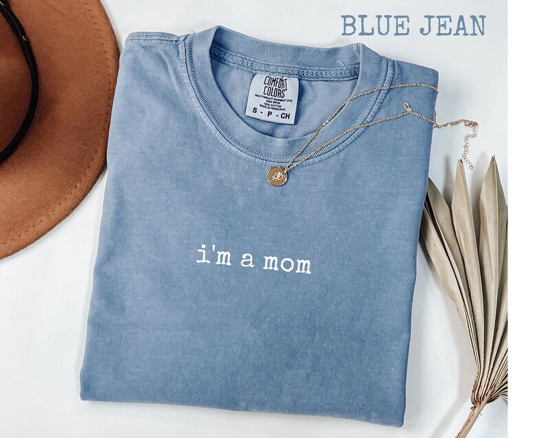 May include: A blue jean Comfort Colors t-shirt with the embroidered text "i'm a mom". A gold necklace with a pendant is draped across the shirt. The shirt is folded and displayed on a white surface.