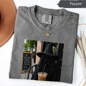 May include: A pepper-coloured crew neck t-shirt featuring a graphic print of a person wearing a black witch hat and sunglasses, holding an iced coffee. The graphic includes a sign that reads Witches Brew Better Coffee. A gold necklace rests on the shirt.