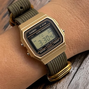 May include: A gold-toned Casio F-91W digital watch with a green nylon strap. The watch face displays the time, day, and date. The watch is water-resistant and has an alarm and chronograph functions.