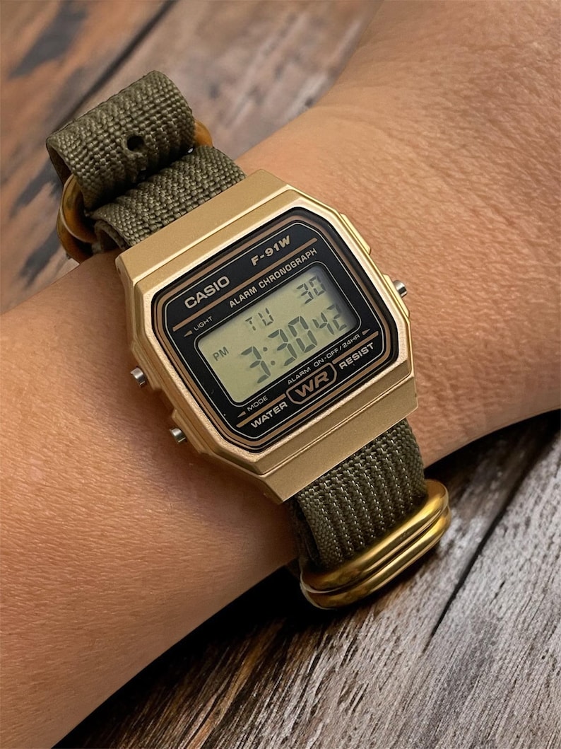 May include: A gold-toned Casio F-91W digital watch with a green nylon strap. The watch face displays the time, day, and date. The watch is water-resistant and has an alarm and chronograph functions.