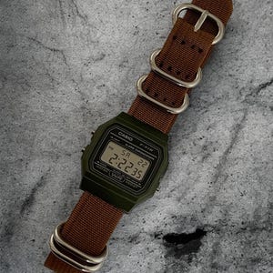 May include: A digital watch with a green case and a brown nylon strap. The watch face displays the time as 14:22:35. The strap has silver-coloured metal hardware.