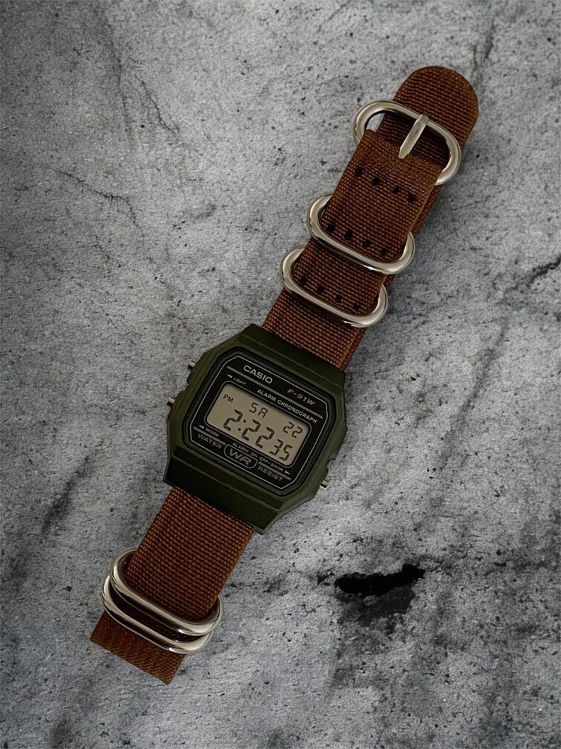 May include: A digital watch with a green case and a brown nylon strap. The watch face displays the time as 14:22:35. The strap has silver-coloured metal hardware.