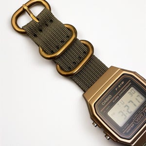 May include: A gold-toned Casio F-91W digital watch with a green nylon strap and gold-colored metal hardware. The watch face displays the time and date. The strap has a classic military style.