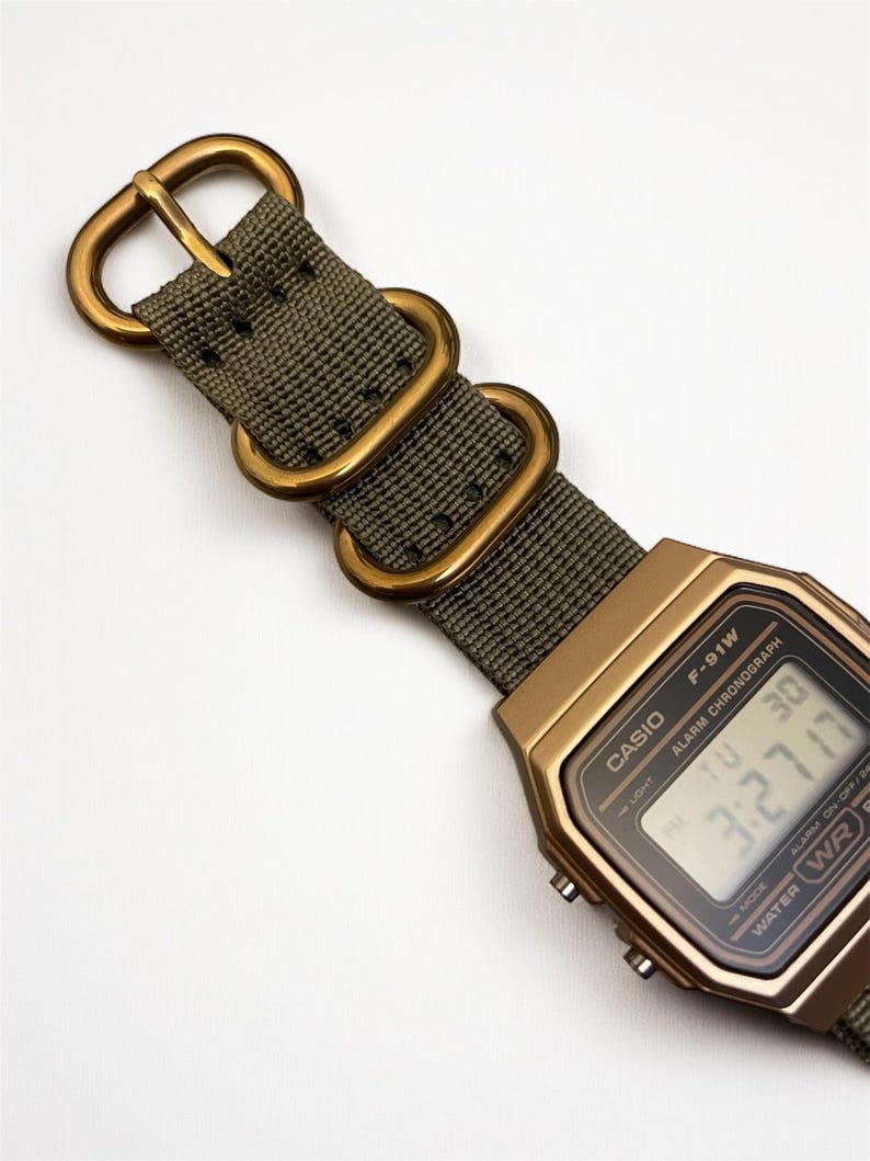 May include: A gold-toned Casio F-91W digital watch with a green nylon strap and gold-colored metal hardware. The watch face displays the time and date. The strap has a classic military style.