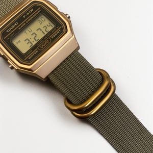 May include: A gold-toned Casio F-91W digital watch with a green fabric strap. The watch face displays the time and date. The strap features gold-colored metal loops. The watch is water-resistant.