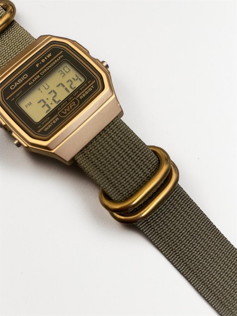 May include: A gold-toned Casio F-91W digital watch with a green fabric strap. The watch face displays the time and date. The strap features gold-colored metal loops. The watch is water-resistant.