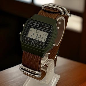 May include: A digital Casio F-91W watch with a dark green case and a brown fabric strap. The watch displays the time as 2:32:25 PM. The watch is displayed on a clear stand.