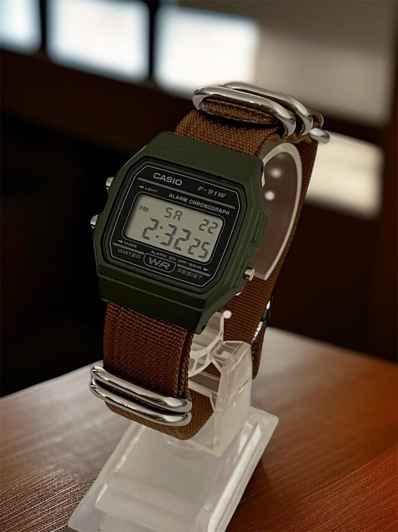 May include: A digital Casio F-91W watch with a dark green case and a brown fabric strap. The watch displays the time as 2:32:25 PM. The watch is displayed on a clear stand.