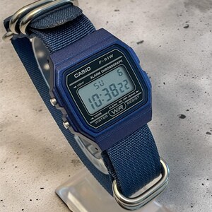 Puede incluir: Un reloj digital Casio F-91W azul con una correa de nailon azul a juego y herrajes de metal plateado. La esfera rectangular muestra la hora 10:38:22, el d&iacute;a SU y la fecha 6, con texto que incluye CASIO, ALARM CHRONOGRAPH y WATER WR RESIST.