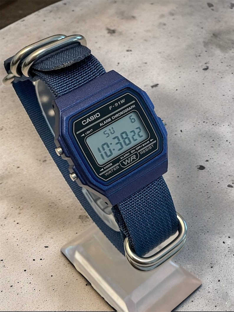 Puede incluir: Un reloj digital Casio F-91W azul con una correa de nailon azul a juego y herrajes de metal plateado. La esfera rectangular muestra la hora 10:38:22, el d&iacute;a SU y la fecha 6, con texto que incluye CASIO, ALARM CHRONOGRAPH y WATER WR RESIST.