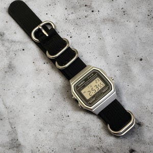 May include: A silver Casio digital watch with a black nylon strap. The watch face displays the time as 2:53:55 PM. The watch has a rectangular face and is water resistant.