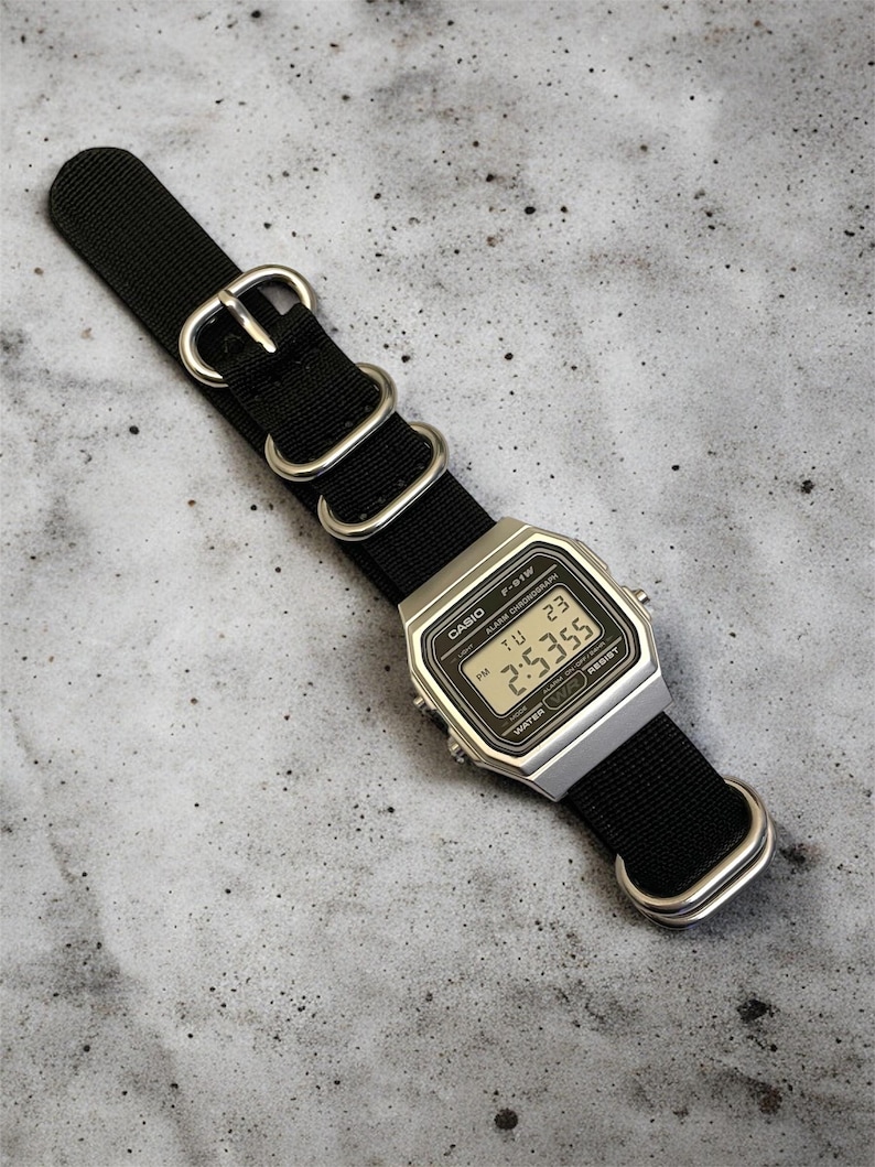 May include: A silver Casio digital watch with a black nylon strap. The watch face displays the time as 2:53:55 PM. The watch has a rectangular face and is water resistant.