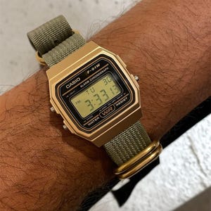 May include: A gold-toned Casio F-91W digital watch with a green fabric band. The watch face displays the time 3:33:13. The watch has a rectangular case and features the Casio logo.