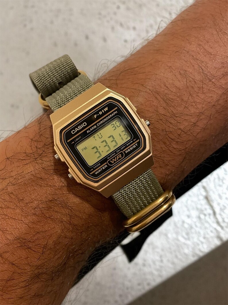 May include: A gold-toned Casio F-91W digital watch with a green fabric band. The watch face displays the time 3:33:13. The watch has a rectangular case and features the Casio logo.