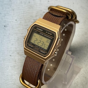 Może przedstawiać: Złoty zegarek cyfrowy Casio F-91W z brązowym paskiem z tkaniny. Tarcza zegarka wyświetla godzinę, dzień i datę. Zegarek ma prostokątną tarczę i złote elementy.