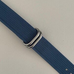 Puede incluir: Una correa de reloj de tela tejida azul con dos anillos rectangulares de metal plateado pulido. La correa tiene una superficie texturizada e incluye dos peque&ntilde;os orificios circulares visibles en el extremo inferior de la banda.
