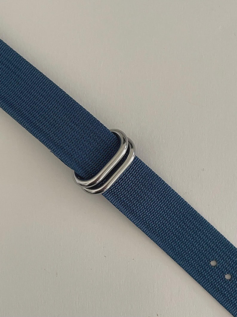 Puede incluir: Una correa de reloj de tela tejida azul con dos anillos rectangulares de metal plateado pulido. La correa tiene una superficie texturizada e incluye dos peque&ntilde;os orificios circulares visibles en el extremo inferior de la banda.
