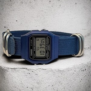 Puede incluir: Un reloj de pulsera digital azul con esfera rectangular y una correa de tela azul a juego, asegurada por dos anillos met&aacute;licos plateados. La pantalla muestra el texto CASIO, F-91W, ALARM CHRONOGRAPH, WATER WR RESIST y la hora 10:39:05.