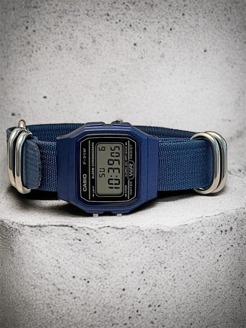 Puede incluir: Un reloj de pulsera digital azul con esfera rectangular y una correa de tela azul a juego, asegurada por dos anillos met&aacute;licos plateados. La pantalla muestra el texto CASIO, F-91W, ALARM CHRONOGRAPH, WATER WR RESIST y la hora 10:39:05.