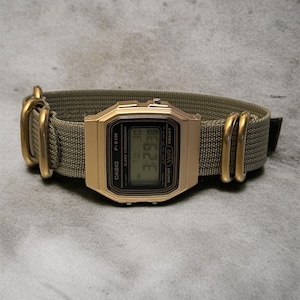 May include: A gold-toned Casio F-91W digital watch with a green nylon strap. The watch face displays the time and date. The strap has gold-colored metal hardware. The watch is displayed on a gray surface.