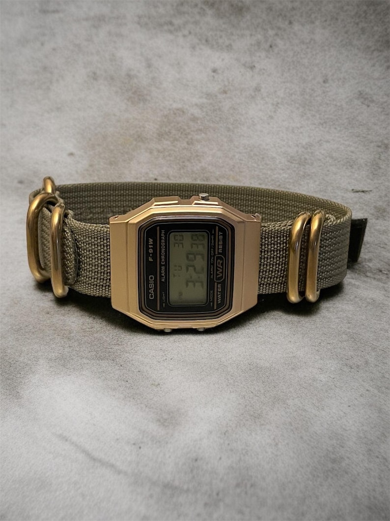 May include: A gold-toned Casio F-91W digital watch with a green nylon strap. The watch face displays the time and date. The strap has gold-colored metal hardware. The watch is displayed on a gray surface.