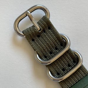 May include: Close-up of an olive green watch strap with silver-colored metal hardware. The strap has a textured weave and multiple metal loops and a buckle. The watch face is partially visible.