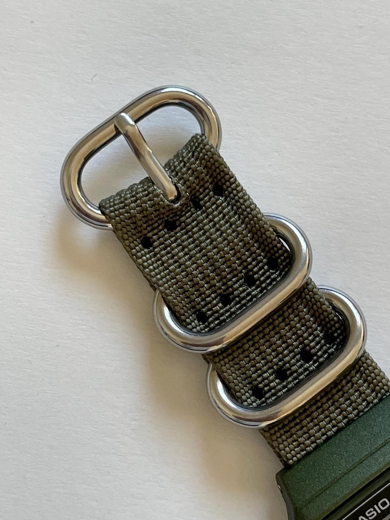 May include: Close-up of an olive green watch strap with silver-colored metal hardware. The strap has a textured weave and multiple metal loops and a buckle. The watch face is partially visible.