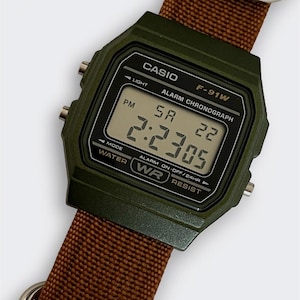 May include: A digital Casio F-91W watch with a dark green case and a brown nylon strap. The watch face displays the time as 2:23:05 PM. The watch has a light, alarm, and chronograph functions.